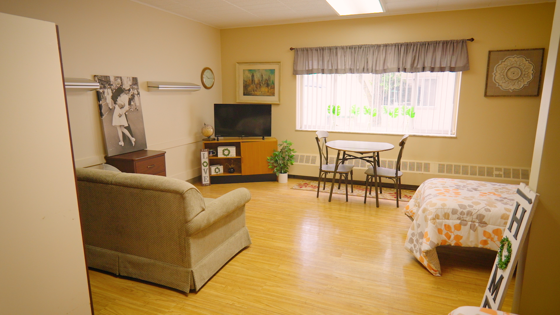 Renovated studio-style resident room at Cedar Woods Assisted Living featuring a cozy seating area, dining table, natural light, and comfortable bed.