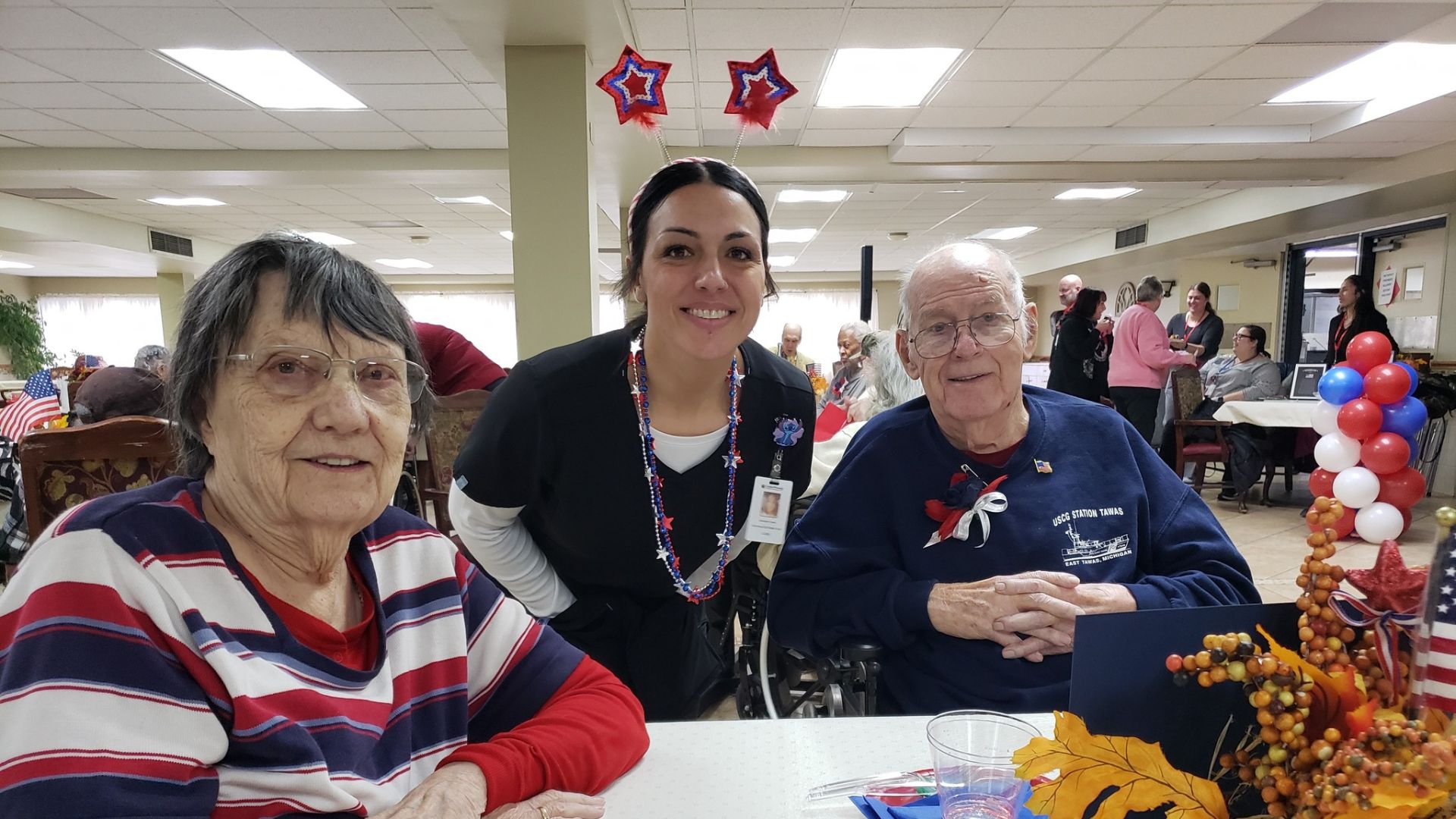 Caregiver smiling with two senior residents during a patriotic Veterans Day celebration at Cedar Woods Assisted Living in Belleville, Michigan